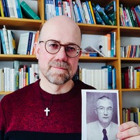 Ein Mann steht vor einem Bücherregeal, er trägt einen rot-schwarzen Pullover, eine Brille, Dreitagebart und eine Glatze, in der Hand hält er ein schwarz-weiß Foto seines Opas 