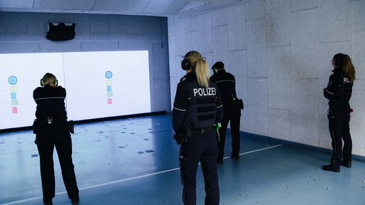Lucie Piwek und Ronican Sansar stehen gebückt, halten eine Pistole vor sich und zielen auf eine Wand. Zwei Ausbilder der Polizei beobachten sie dabei.