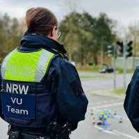 Zwei Polizisten stehen mit dem Rücken zur Kamera. Sie tragen jeweils eine Weste mit der Aufschrift "Polizei NRW VU Team".