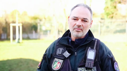 Sascha Mill steht in seiner Polizeiuniform der Kamera zugewendet, hinter ihm ist eine Wiese unscharf zu erkennen.