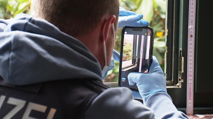 Ermittler Julius Mergelkamp fotografiert mit einem Handy die Schäden an einem Holzfensterrahmen mit Einbruchsspuren