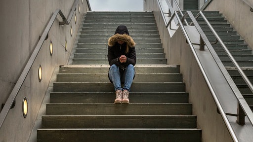 Eine Jugendliche sitzt mit gesenktem Kopf auf einer Treppe, sie trägt eine Kaputze.
