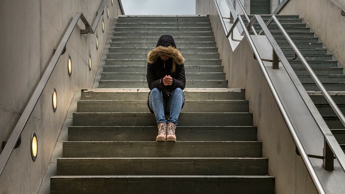 Eine Jugendliche sitzt mit gesenktem Kopf auf einer Treppe, sie trägt eine Kaputze.