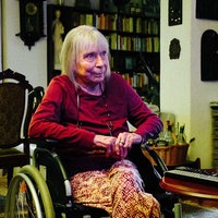 Renate Nowak sitzt in einem Rollstuhl im Wohnzimmer und schaut Fernsehen.
