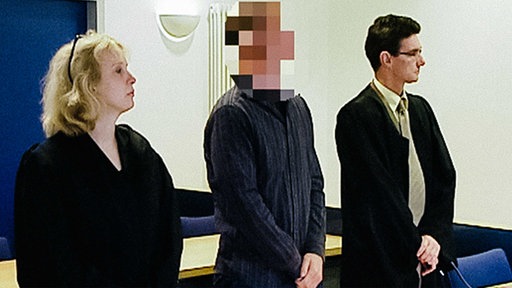 Eine Frau und zwei Männer stehen vor einem Tisch, die Frau steht links, in der Mitte der Verdächtige mit gepixeltem Gesicht, rechts daneben der andere Mann, sie tragen alle dunkle Kleidung