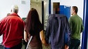 Drei Männer und eine Frau von hinten, die Frau hält die Hand des Mannes, der Handschellen trägt, sein Hinterkopf ist verpixelt 
