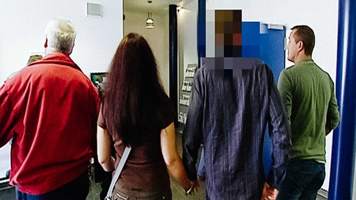 Drei Männer und eine Frau von hinten, die Frau hält die Hand des Mannes, der Handschellen trägt, sein Hinterkopf ist verpixelt 