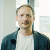 Ein Mann mit weißem T-Shirt und grauer Jacke steht in einem Büro, hinter ihm ein Fenster.