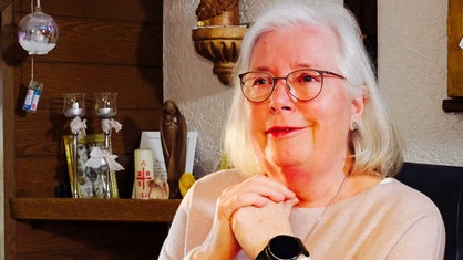 Eine ältere Frau mit kinnlangen weißen Haaren und Brille sitzt in einer Wohnstube 