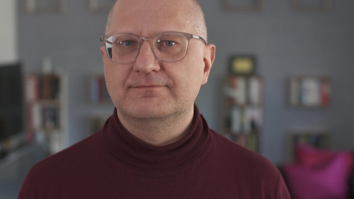 Mann mit transparenter Brille und weinrotem Rolli schaut in Kamera 