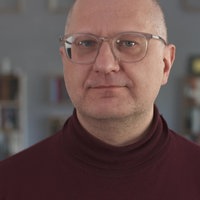 Mann mit transparenter Brille und weinrotem Rolli schaut in Kamera 