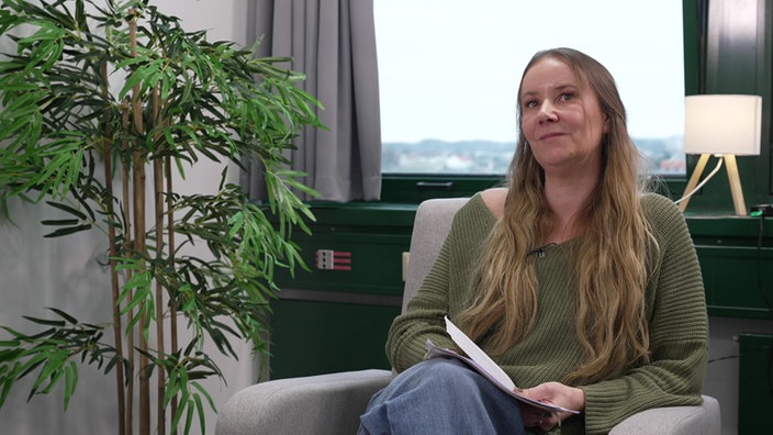 Kriminaloberkommissarin Maike Schmidt sitzt auf einem Stuhl vor einem Fenster und hält eine Akte in den Händen.
