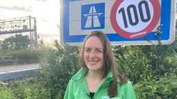 Eine Frau mit Greenpeace-Jacke steht vor einem Autobahnschild