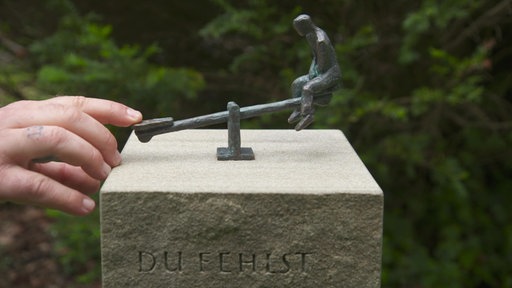 Ein Finger drückt eine Messingfigur auf einer Wippe in die Luft