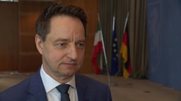 Man sieht einen Mann in Anzug, im Hintergrund sind Flaggen zu sehen.