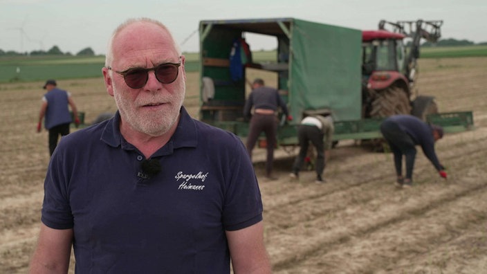 Ein Mann mit Sonnenbrille steht auf einem Feld vor einem Traktor. Daneben ernten vier Männer Spargel