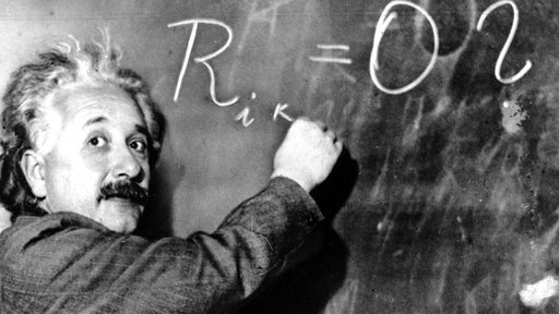 Ein Schwarz-Weiß-Foto zeigt Albert Einstein, wie er eine Formel mit Kreide an eine Tafel schreibt und in die Kamera blickt