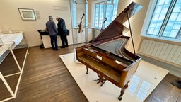 Beethovens letzter Flügel steht auf einem Podest im Bonner Beethoven-Haus, im Hintergund stehen zwei Männer.