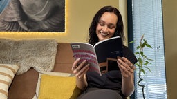 Myra Abel sitzt auf einer Couch und liest in einem ihre Gedichtbände, das sie in der Hand hält