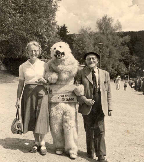 Ein Mensch im Eisbärkostüm hat sich bei einem Mann und einer Frau eingehakt. Auf einem Schild steht "Gruß vom Titisee".