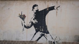 Das Bild zeigt ein Graffiti: Ein vermummter Mann wirft einen Blumenstrauß