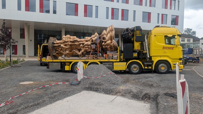 Ein gelber Lkw steht vor einem weißen Haus. Auf der Ladefläche ist eine bronzefarbene Skulptur befestigt. Im Vordergrund liegt Schotter. 