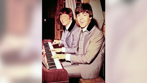 Paul McCartney und John Lennon sitzen gemeinsam am Klavier und lächeln Richtung Kamera (ca. 1964)