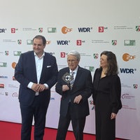 Frank Elstner hält seinen Ehrenpreis in die Kamera. Links neben ihm steht NRW-Medienstaatssekretär Nathanael Liminski, rechts die Grimme-Geschäftsführerin Çiğdem Uzunoğlu.