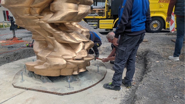 Auf einem Beton-Fundament steht eine bronzene Statue. Handwerker knien davor und schlagen mit einem Hammer silberne Gewindestangen in die Plattform auf der die Skulptur steht, um sie auf dem Fundament zu befestigen. 