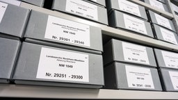 Archivkartons mit Akten des Entnazifizierungshauptausschusses des Regierungsbezirks Köln in einem Regal.