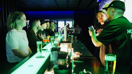 Mehrere Menschen sitzen am Tresen einer Bar, auf der anderen Seite zwei Männer, die mit Gläsern hantieren