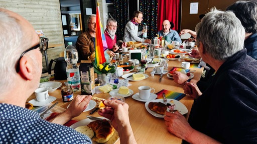 Senioren sitzen um einen gedeckten Frühstückstisch herum. Auf dem Tisch steht eine Regenbogenflagge.
