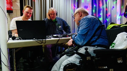 Drei Männer sitzen am DJ-Pult auf der inklusiven Party, einer von ihnen im Rollstuhl
