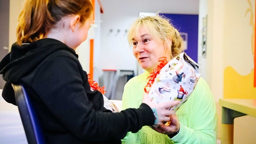 Susanne Saage mit grellgelbem Pullover, hellblonden Haaren, kniet vor einem Mädchen mit braunen Haaren und schwarzem Pulli, Saage reicht ihr zwei Geschenke