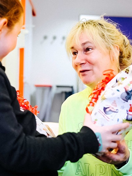 Susanne Saage mit grellgelbem Pullover, hellblonden Haaren, kniet vor einem Mädchen mit braunen Haaren und schwarzem Pulli, Saage reicht ihr zwei Geschenke