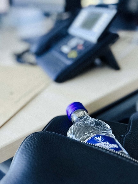 Eine kleine Vodkaflasche ragt aus einer Aktentasche hervor. Im Hintergrund erkennt man einen Schreibtisch, auf dem ein Telefon und eine Akte liegt. 