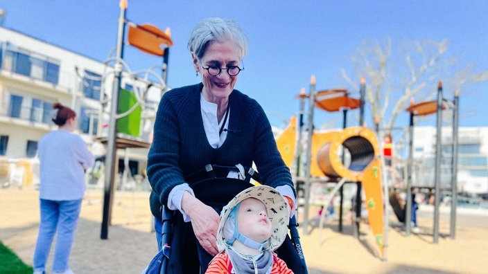 Ein Spielplatz. Katrin Hesse steht hinter einem Kinderwagen. Darin sitzt Philipp. Sie lächelt.