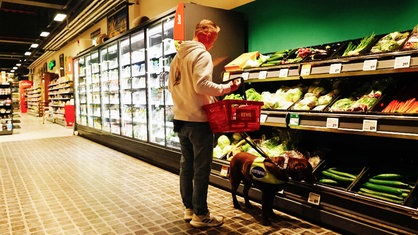 Ein Mann steht vor einem Gemüseregal im Supermarkt. An der Leine hat er einen braunen Labrador.