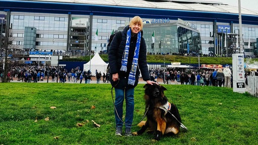 Eine junge Frau steht neben einem Hund vor einem großen Fußballstadion. Vor dem Stadion sind viele Menschen. Einige tragen Fanschals von dem Verein Schalke. Die junge Frau trägt auch einen blauen Fußballschal und legt ihre Hand auf den Kopf des Hundes.