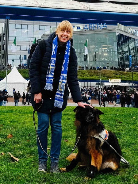 Eine junge Frau steht neben einem Hund vor einem großen Fußballstadion. Vor dem Stadion sind viele Menschen. Einige tragen Fanschals von dem Verein Schalke. Die junge Frau trägt auch einen blauen Fußballschal und legt ihre Hand auf den Kopf des Hundes.