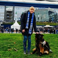Eine junge Frau steht neben einem Hund vor einem großen Fußballstadion. Vor dem Stadion sind viele Menschen. Einige tragen Fanschals von dem Verein Schalke. Die junge Frau trägt auch einen blauen Fußballschal und legt ihre Hand auf den Kopf des Hundes.