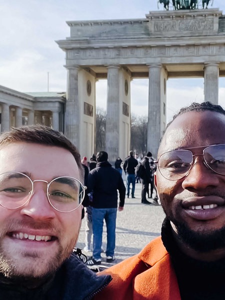 Simon Rieken, weiß, kurze blonde Haare hat einen Arm um Albert Sagno, schwarz, Brille, die schwarzen Haare zurückgebunden, gelegt, im Hintergrund ist das Brandenburger Tor in Berlin zu sehen