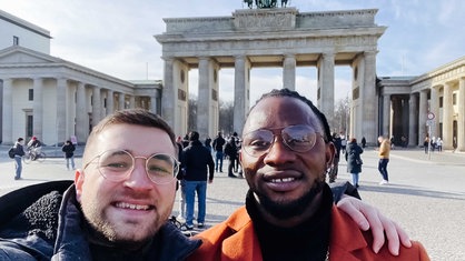 Simon Rieken, weiß, kurze blonde Haare hat einen Arm um Albert Sagno, schwarz, Brille, die schwarzen Haare zurückgebunden, gelegt, im Hintergrund ist das Brandenburger Tor in Berlin zu sehen