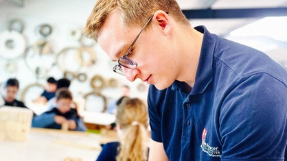 Justus Kötter steht gebeugt über seiner Arbeit. Im Hintergrund werkeln Kinder an Tischen.