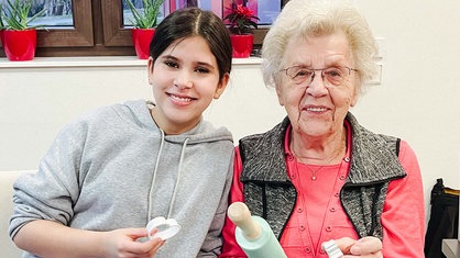 Eine Jugendliche und eine Seniorin lächeln in die Kamera, sie halten eine Keksform und eine Teigrolle in den Händen