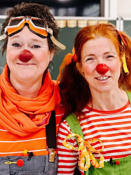 Zwei Frauen sind als Clowns kostümiert und stehen in einem Klassenraum vor einer Tafel.