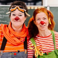 Zwei Frauen sind als Clowns kostümiert und stehen in einem Klassenraum vor einer Tafel.