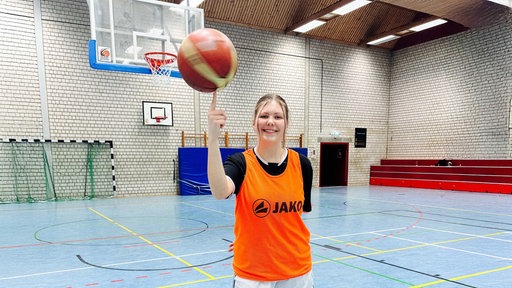 Lilly Schmidt, eine junge Frau in Sportklamotten und einem orangen Leibchen steht in einer Turnhalle und balanciert einen Basketball auf einem Finger.