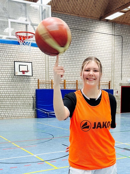Lilly Schmidt, eine junge Frau in Sportklamotten und einem orangen Leibchen steht in einer Turnhalle und balanciert einen Basketball auf einem Finger.