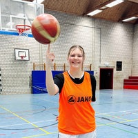 Lilly Schmidt, eine junge Frau in Sportklamotten und einem orangen Leibchen steht in einer Turnhalle und balanciert einen Basketball auf einem Finger.
